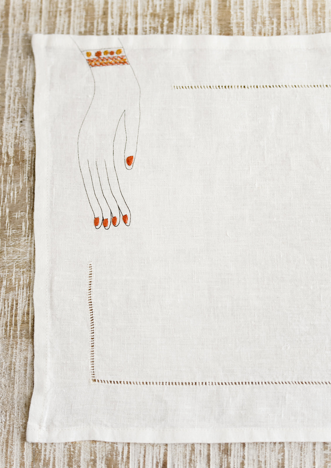 A white linen placemat with hemstitch border and embroidered hand at corner.