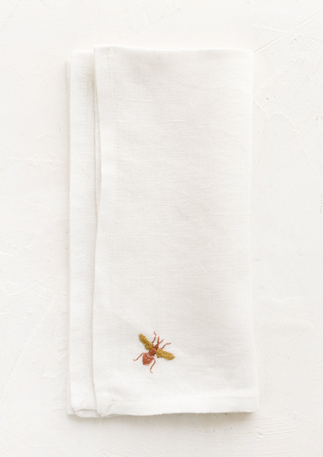 A white linen napkin with single bee embroidery at corner in peach and mustard.