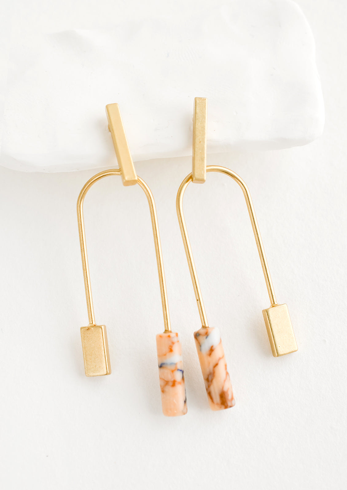 A pair of gold earrings with bar shaped top and arched bottom with peach gemstone on one side.