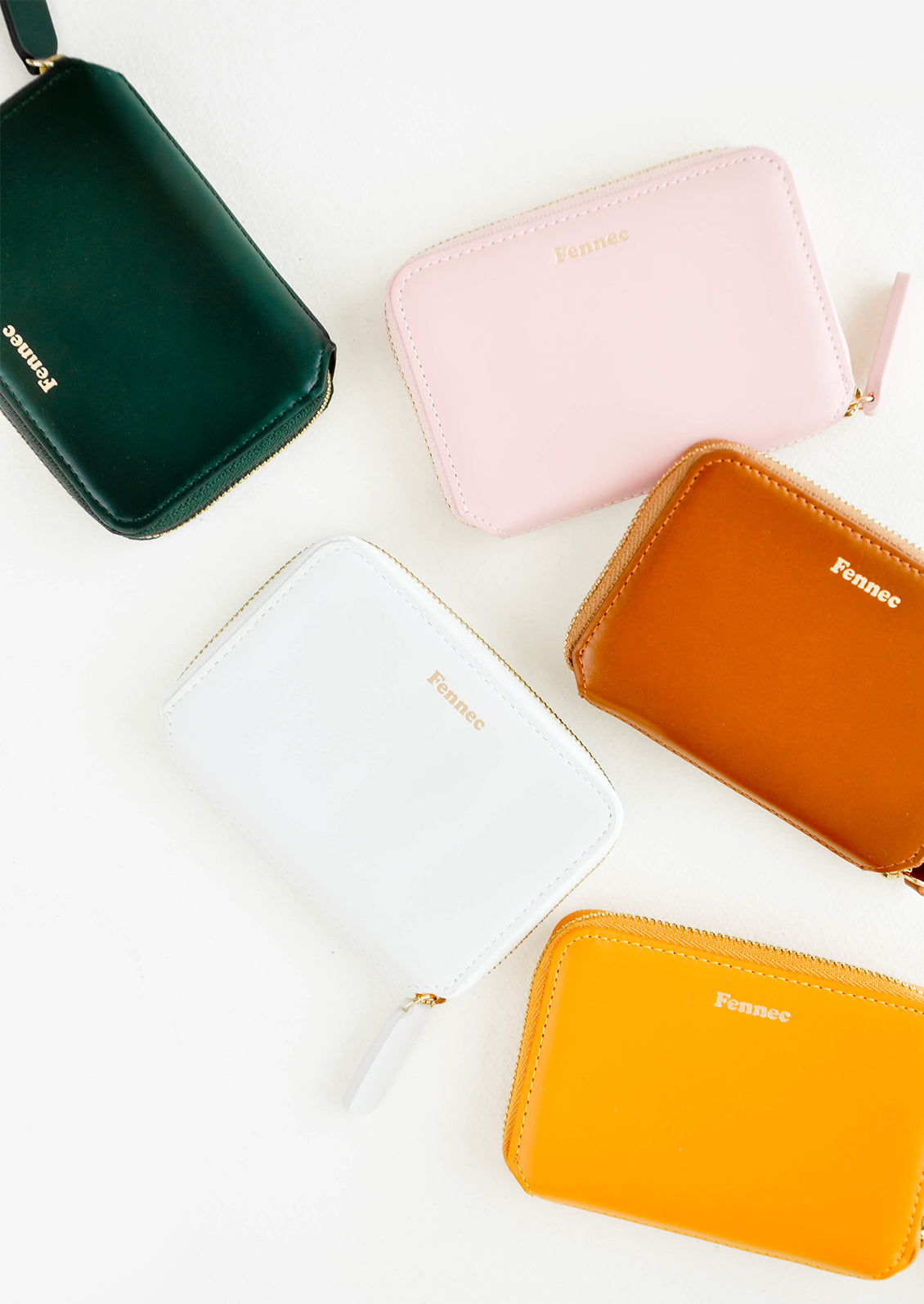 Five small leather zip wallets in dark green, pale pink, rust, mustard yellow, and pale gray all embossed with gold Fennec name. 