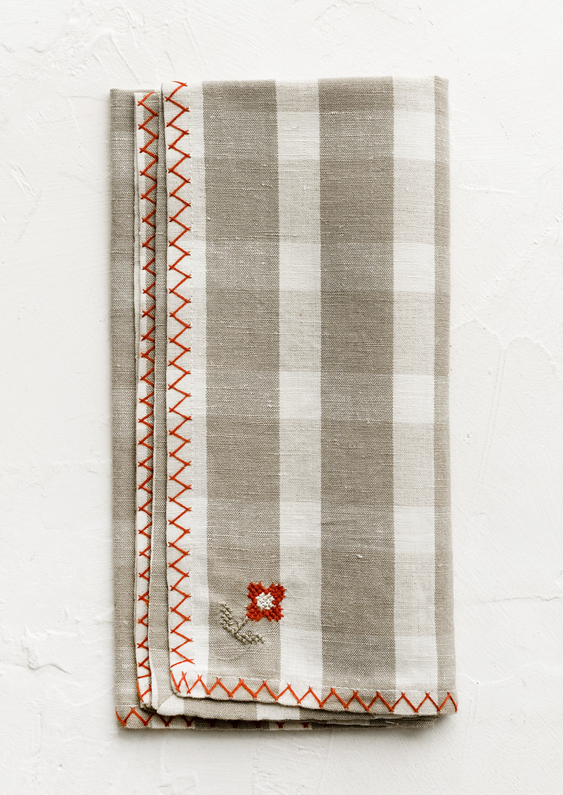 A plaid print napkin in beige and white with red embroidery detailing.