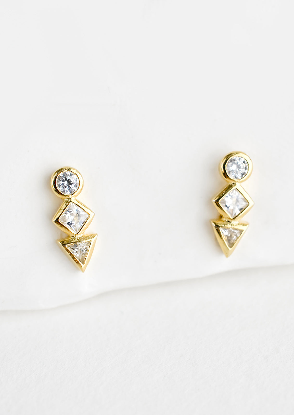 A pair of gold stud earrings with a circle, diamond and triangle crystal stacked in a row.