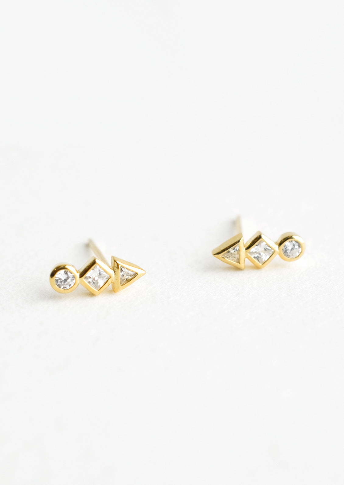A pair of gold stud earrings with a circle, diamond and triangle crystal stacked in a row.