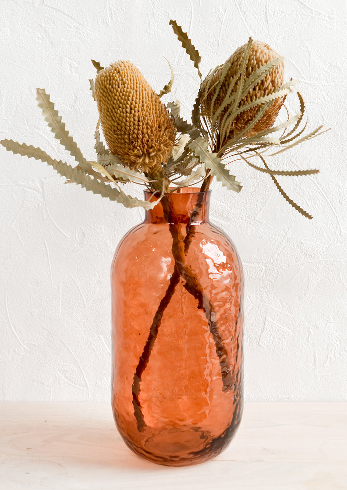 A tall glass vase in rosewood hue with hammered texture, displaying dried banksia flowers.