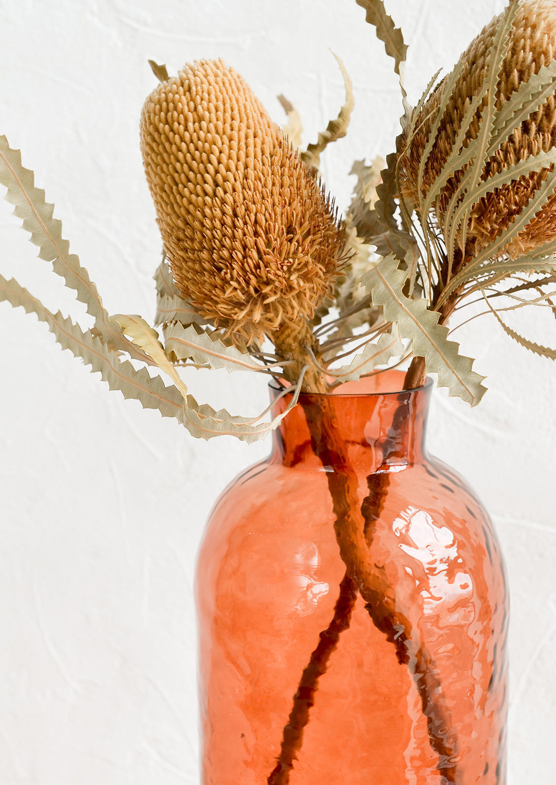 A tall glass vase in rosewood hue with hammered texture, displaying dried banksia flowers.