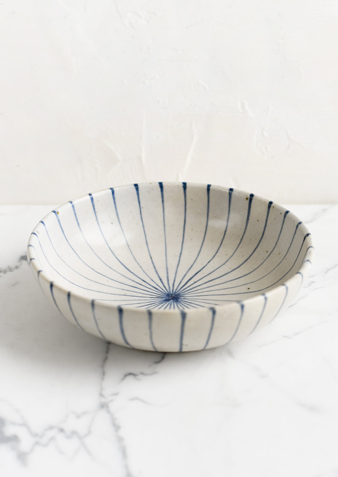 A round and shallow bowl in white with blue sunray pinstripe pattern.