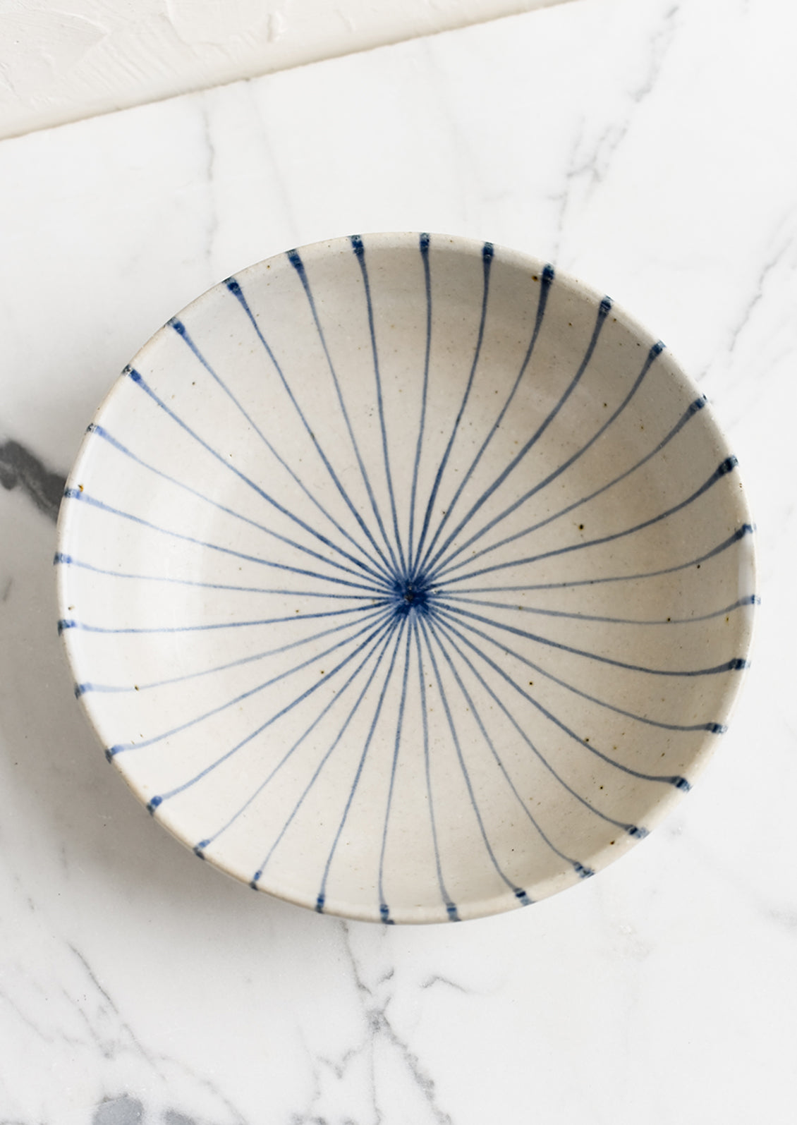 A round and shallow bowl in white with blue sunray pinstripe pattern.
