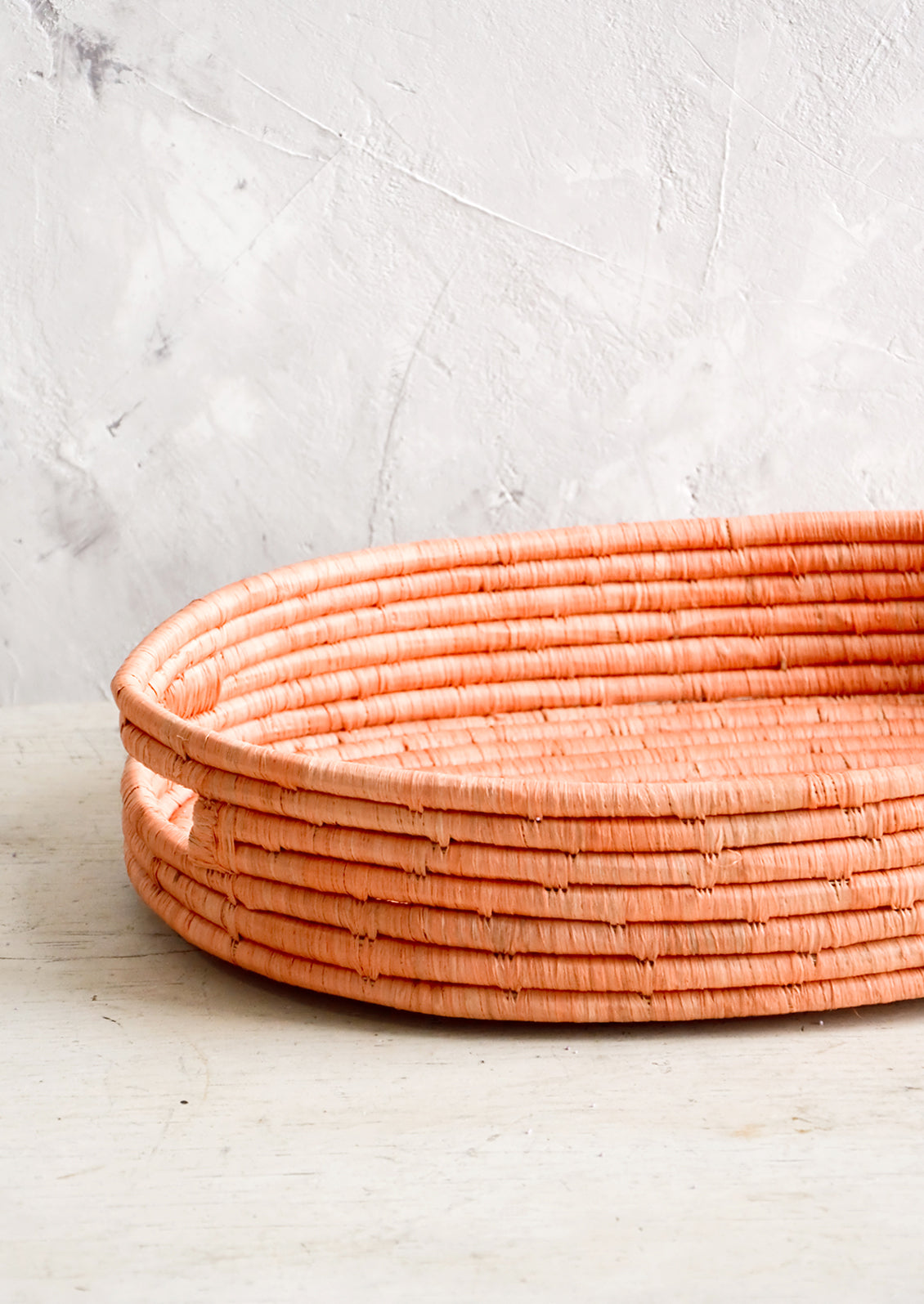 Oval shaped, shallow basket woven in peach raffia with cutout handles at sides.