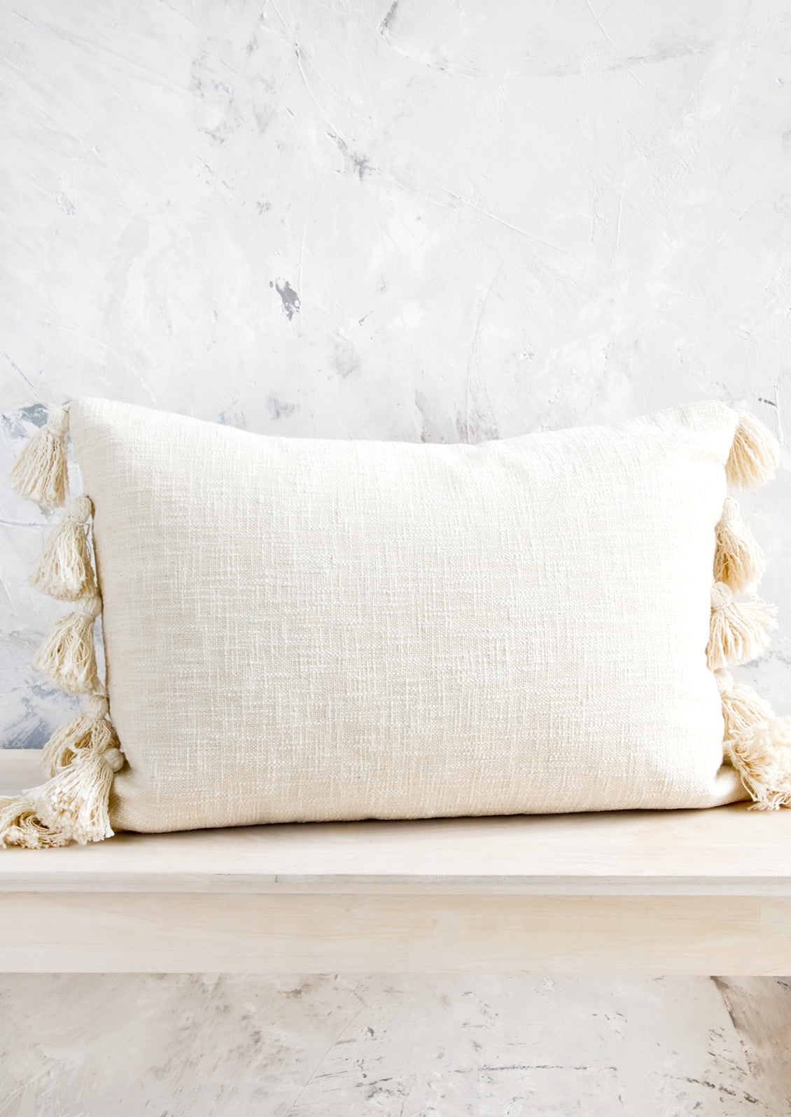 Lumbar throw pillow in textured, cream colored cotton with large decorative tassels at sides.