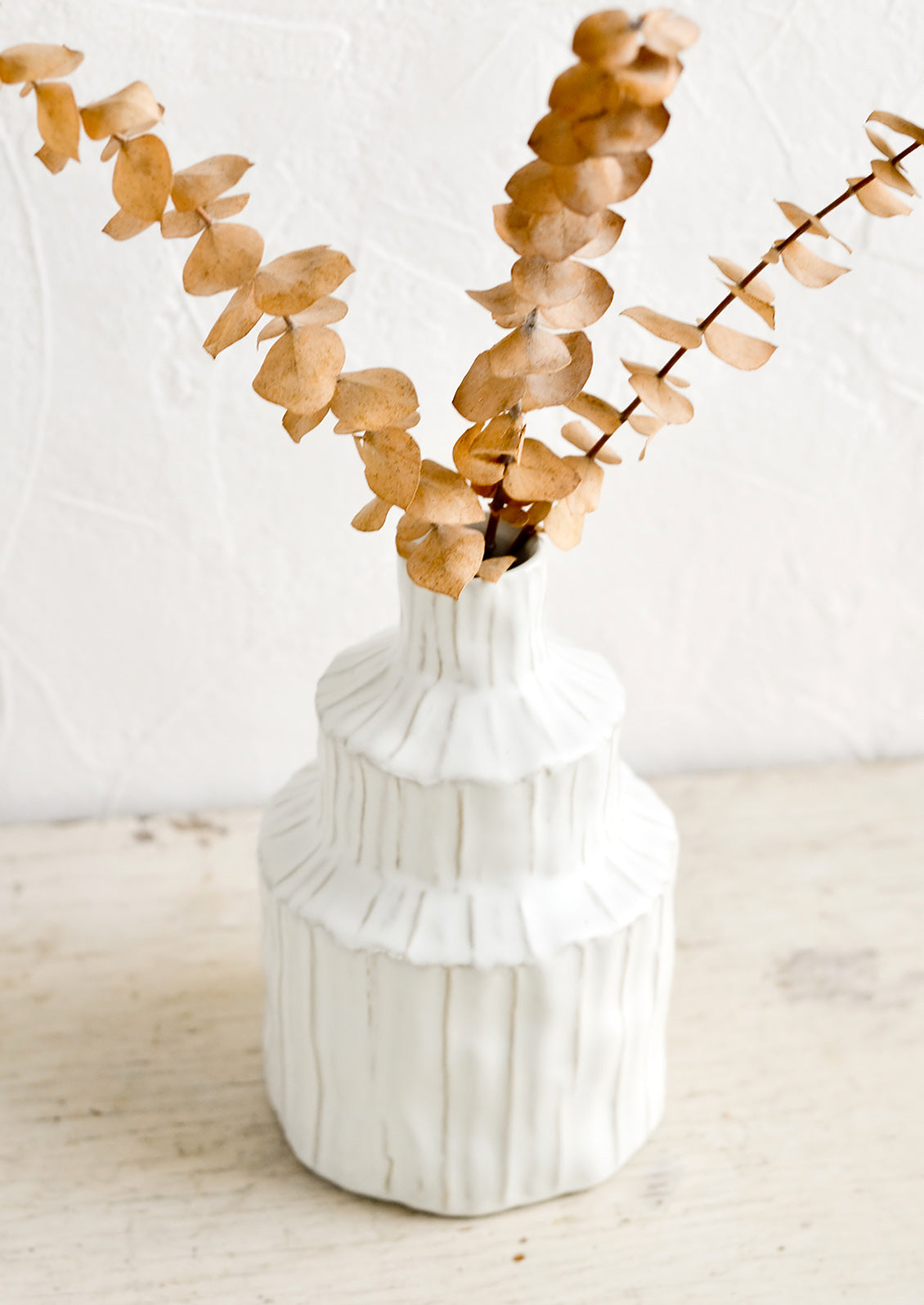 A textured white vase in an asymmetrical shape with dried eucalyptus.