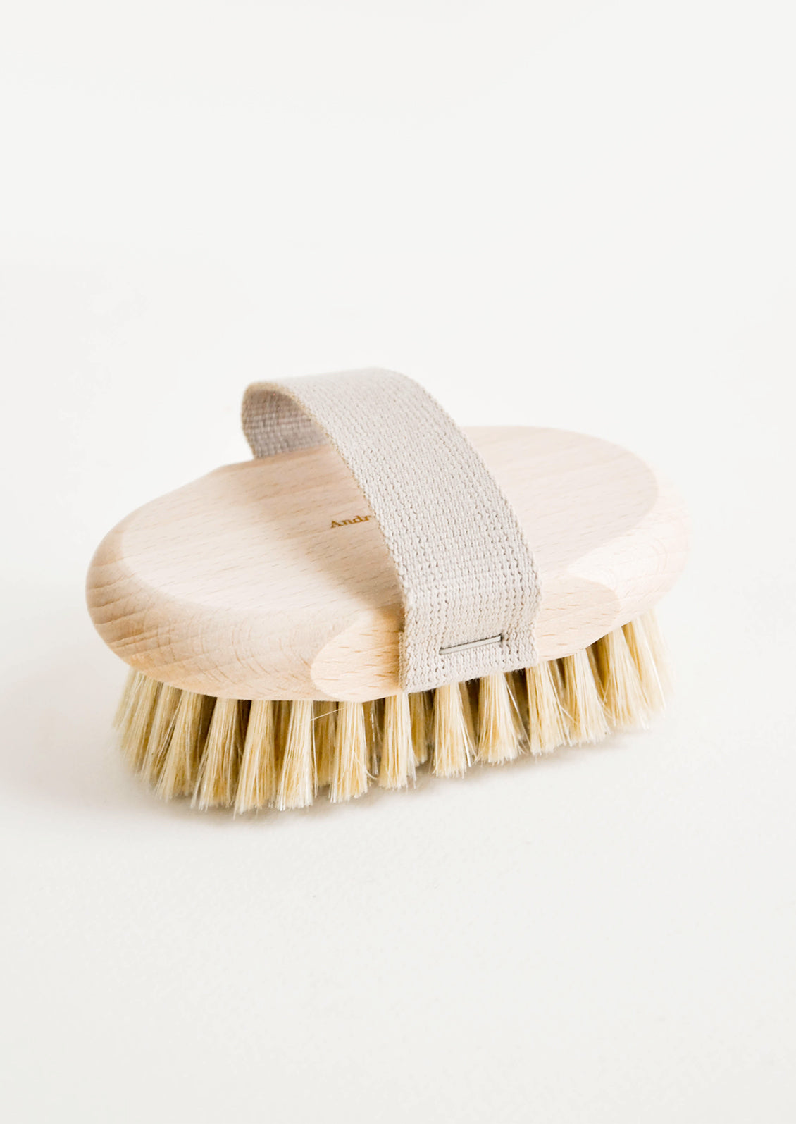 An oval body brush in a light colored natural wood with a cotton hand strap.