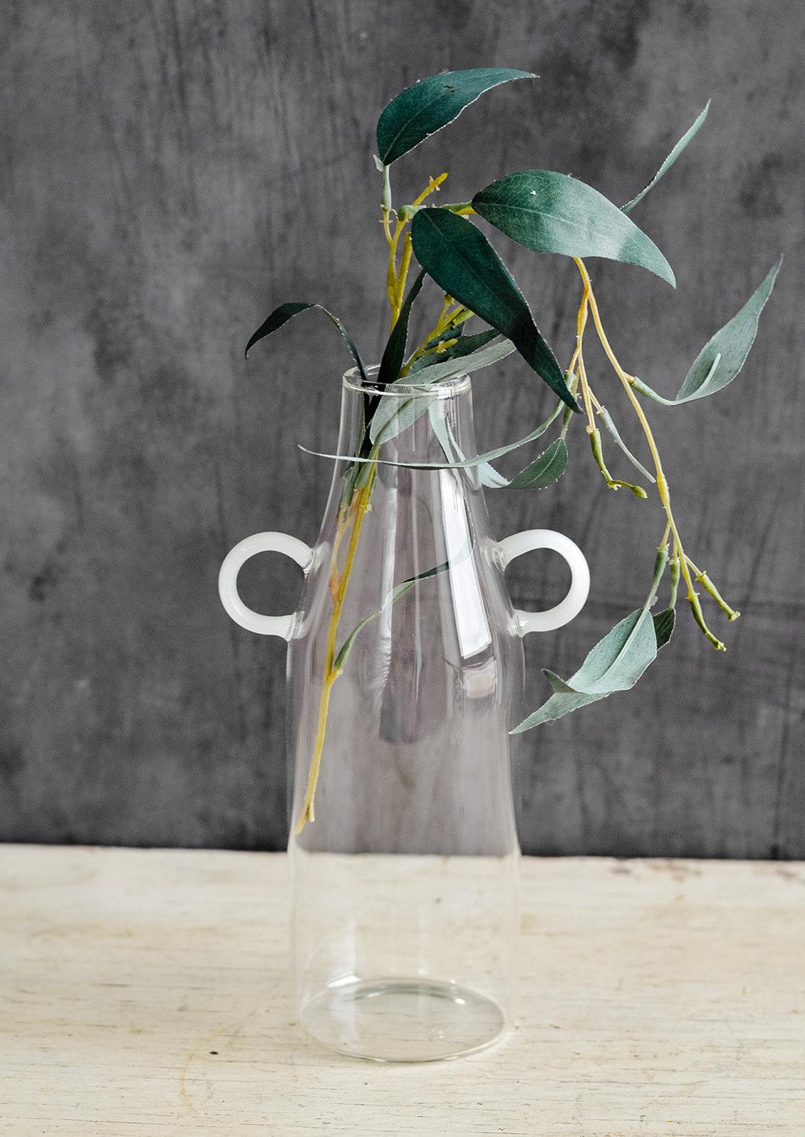 A clear glass vase in bottle shape with curved white handles at sides.