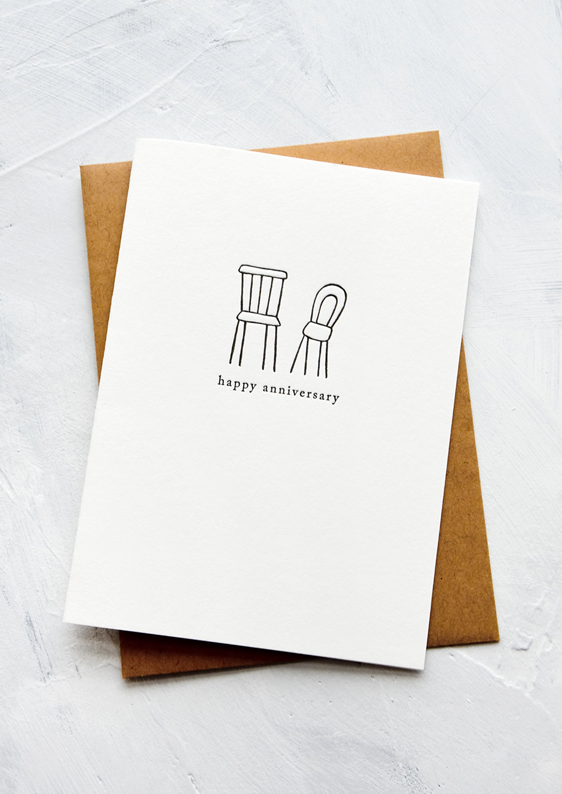 A letterpress printed greeting card with an image of two chairs side by side, text underneath reads "happy anniversary".