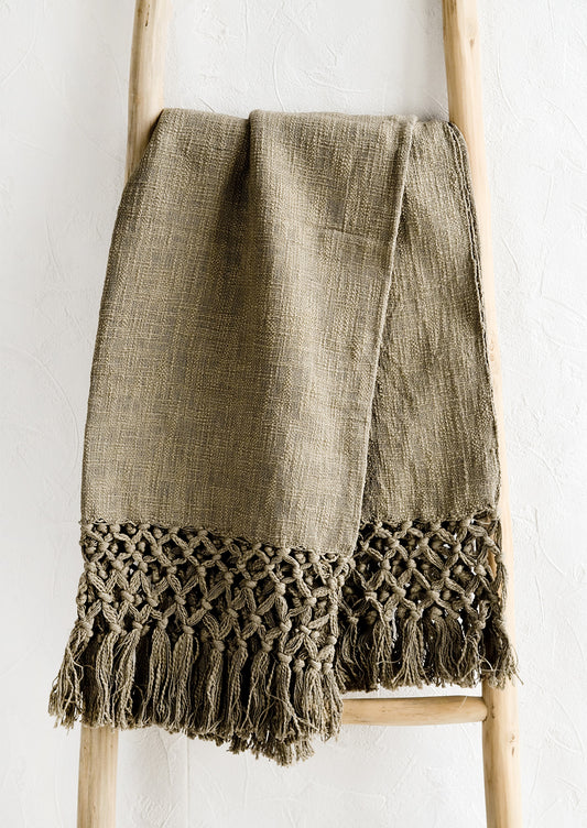 An army green cotton throw blanket with knotted open weave trim, displayed on a ladder.