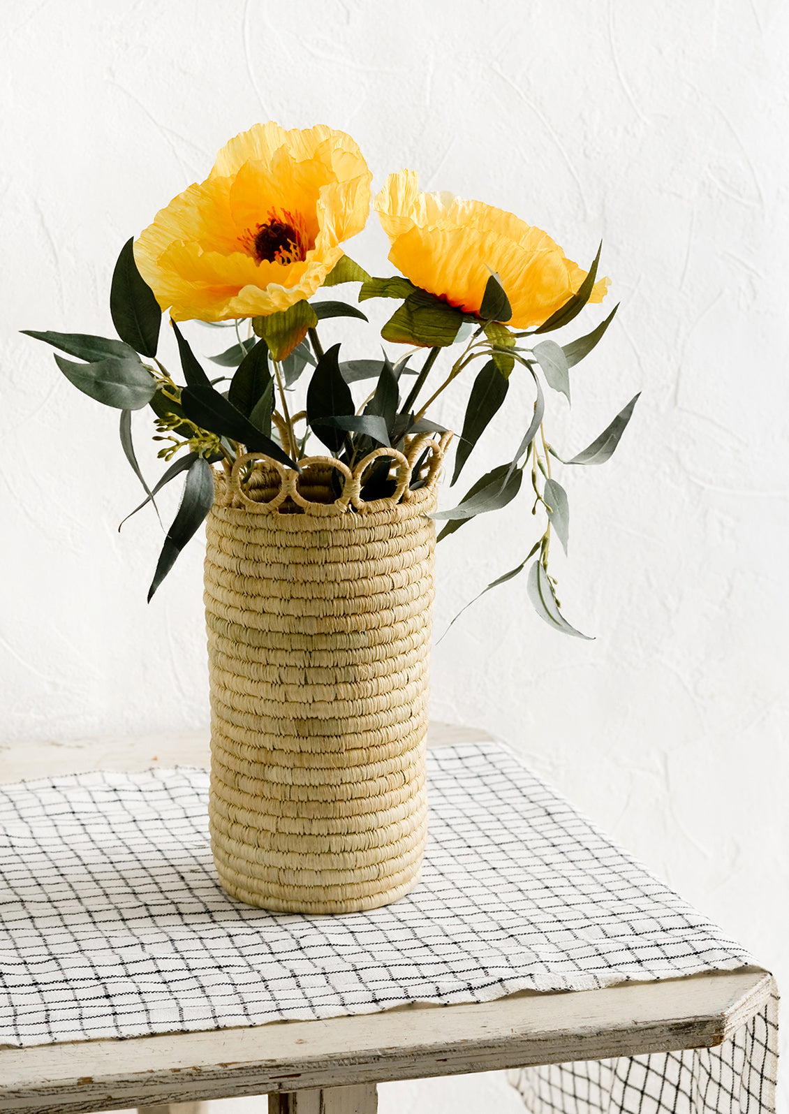 A cylindrical raffia vase on a linen-clad table with eucalyptus and poppies.