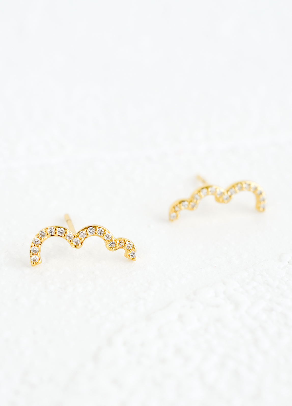A pair of gold stud earrings in squiggly, half cloud-like shape with clear crystal detailing.