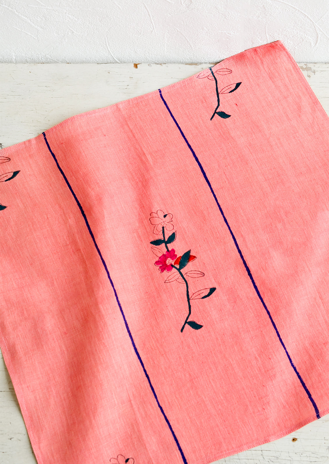 A pink linen napkin with a minimal floral block print design and two vertical blue stripes.
