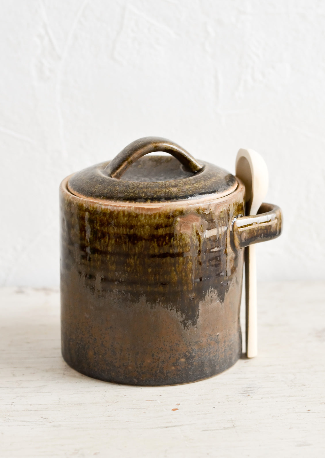 A ceramic storage jar in a rustic dark brown glaze with a wooden spoon on the side.