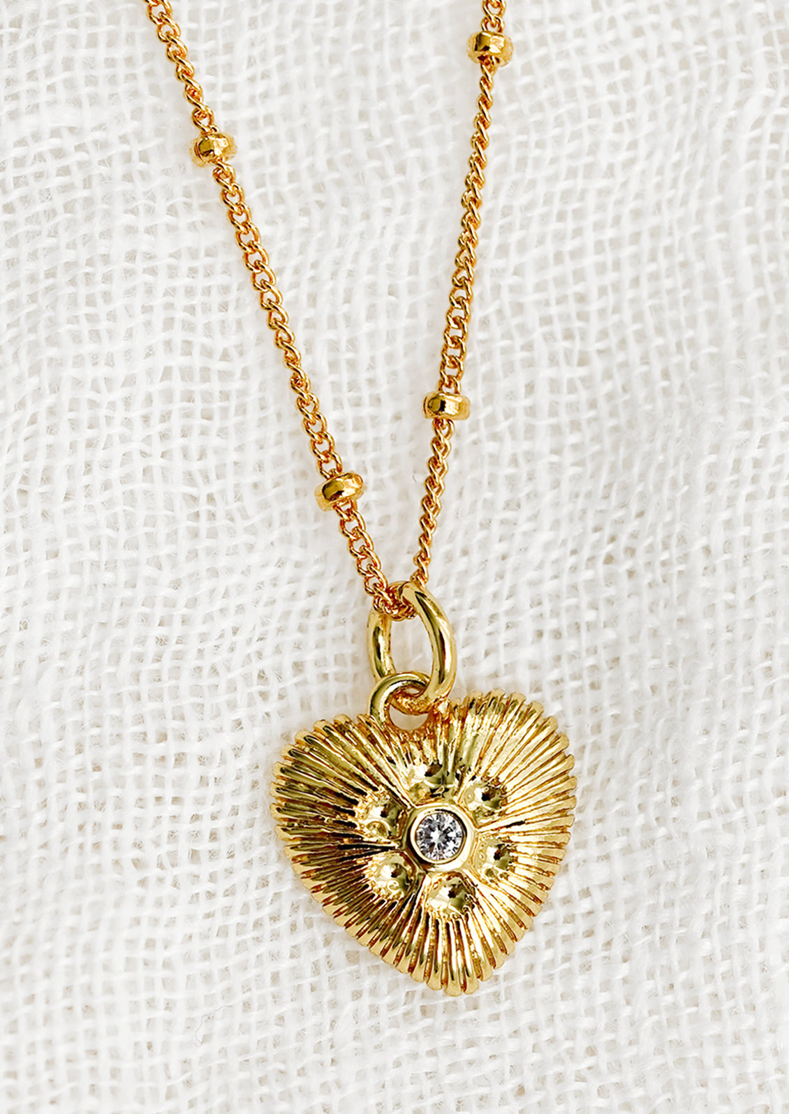A gold necklace with heart shaped charm with ridged texture and crystal detailing.