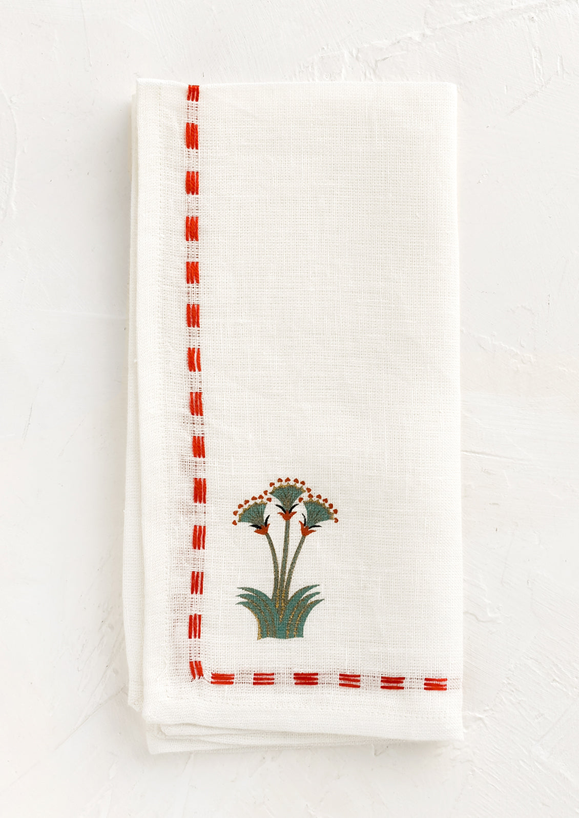 A white linen napkin with stitched red border and nile lotus emblem at corner.
