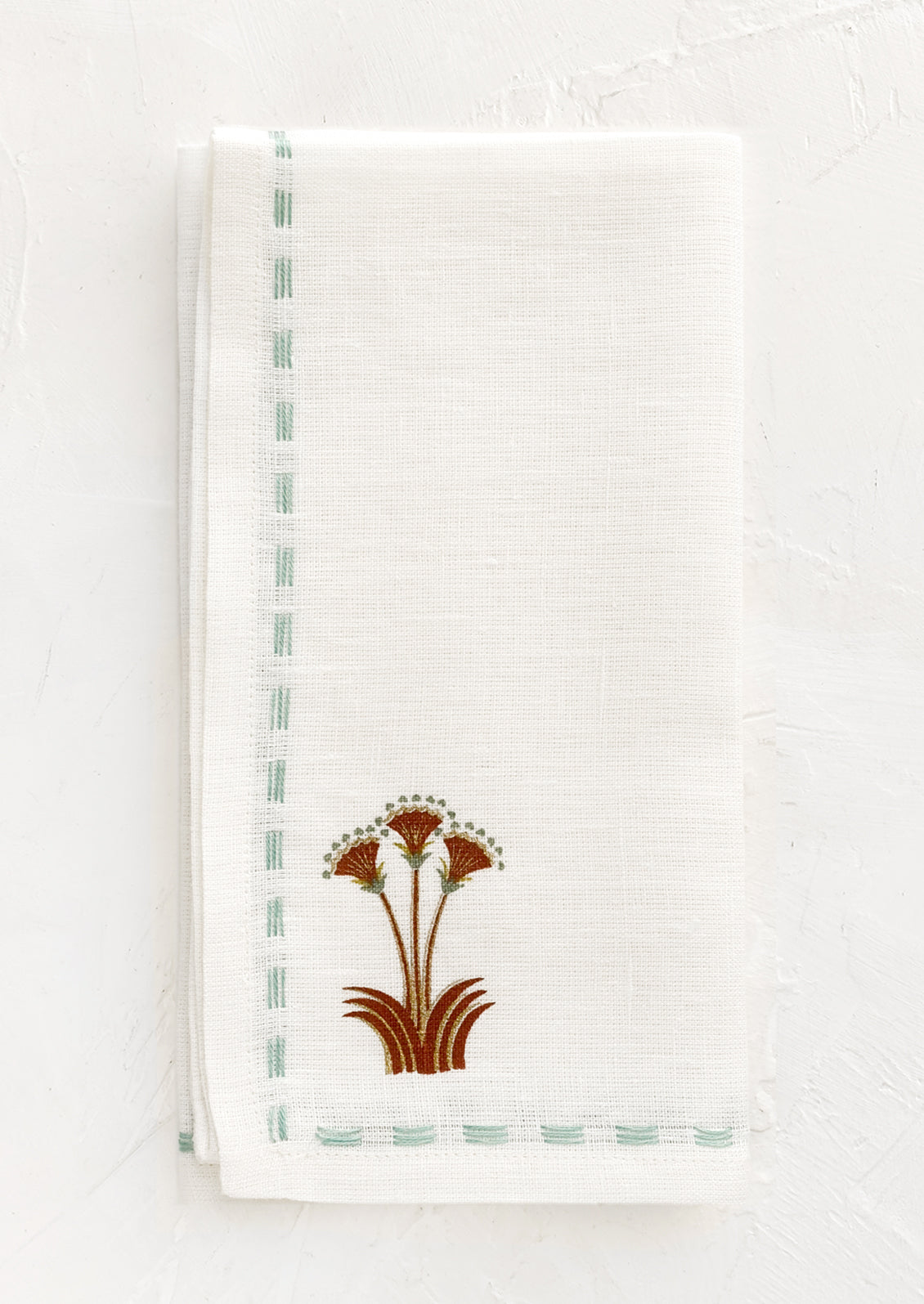 A white linen napkin with stitched seafoam border and nile lotus emblem at corner.