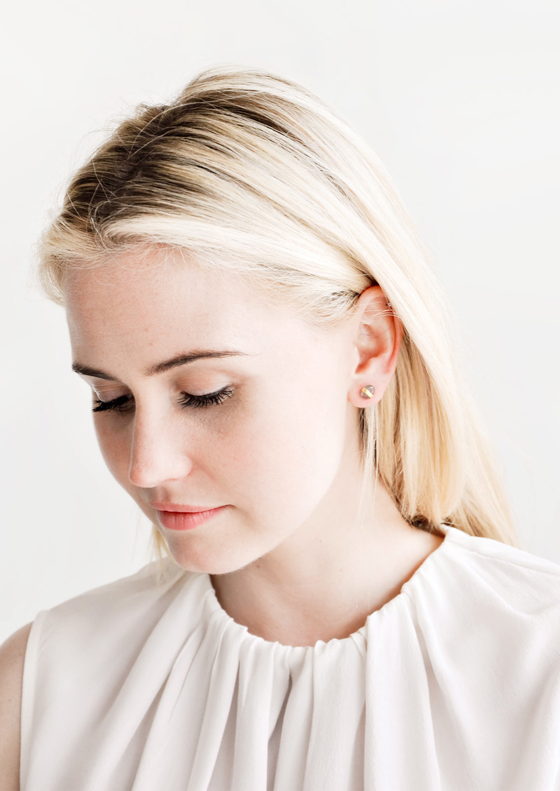 Blonde model looking down, wearing silky white blouse and pebble stud earrings.
