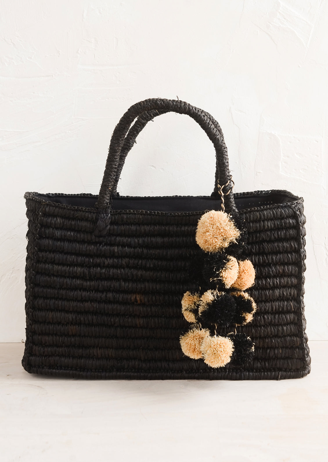 An east-west raffia tote in black with straw pom pom detailing.
