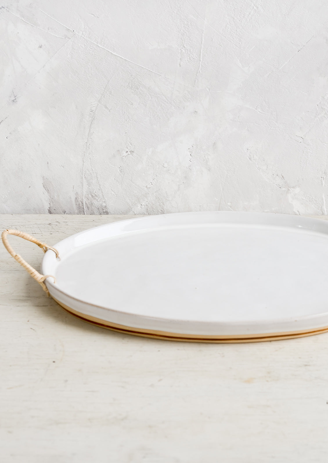 A glossy white ceramic platter in oval shape with rattan wrapped handles at sides.