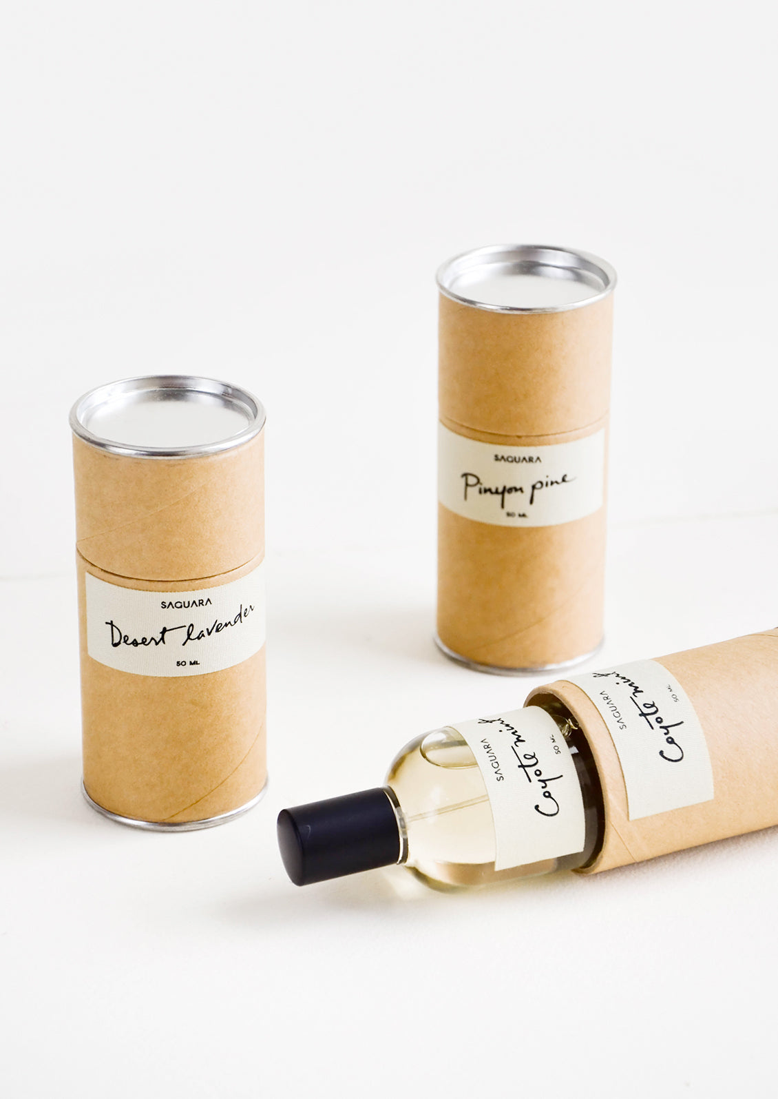 Three cardboard cylinder containers with white labels and black text. One tipped on side with glass perfume bottle inside.
