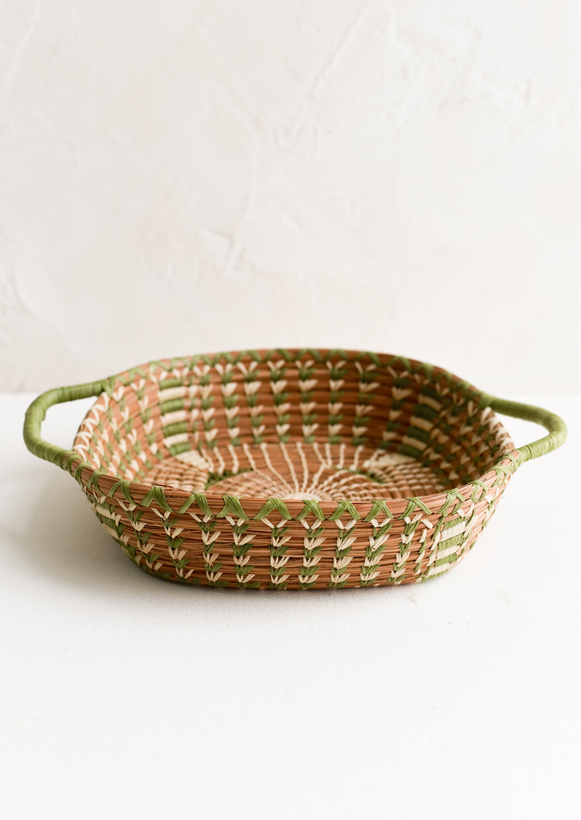 A shallow basket in rounded square shape with rectangular green handles at sides.