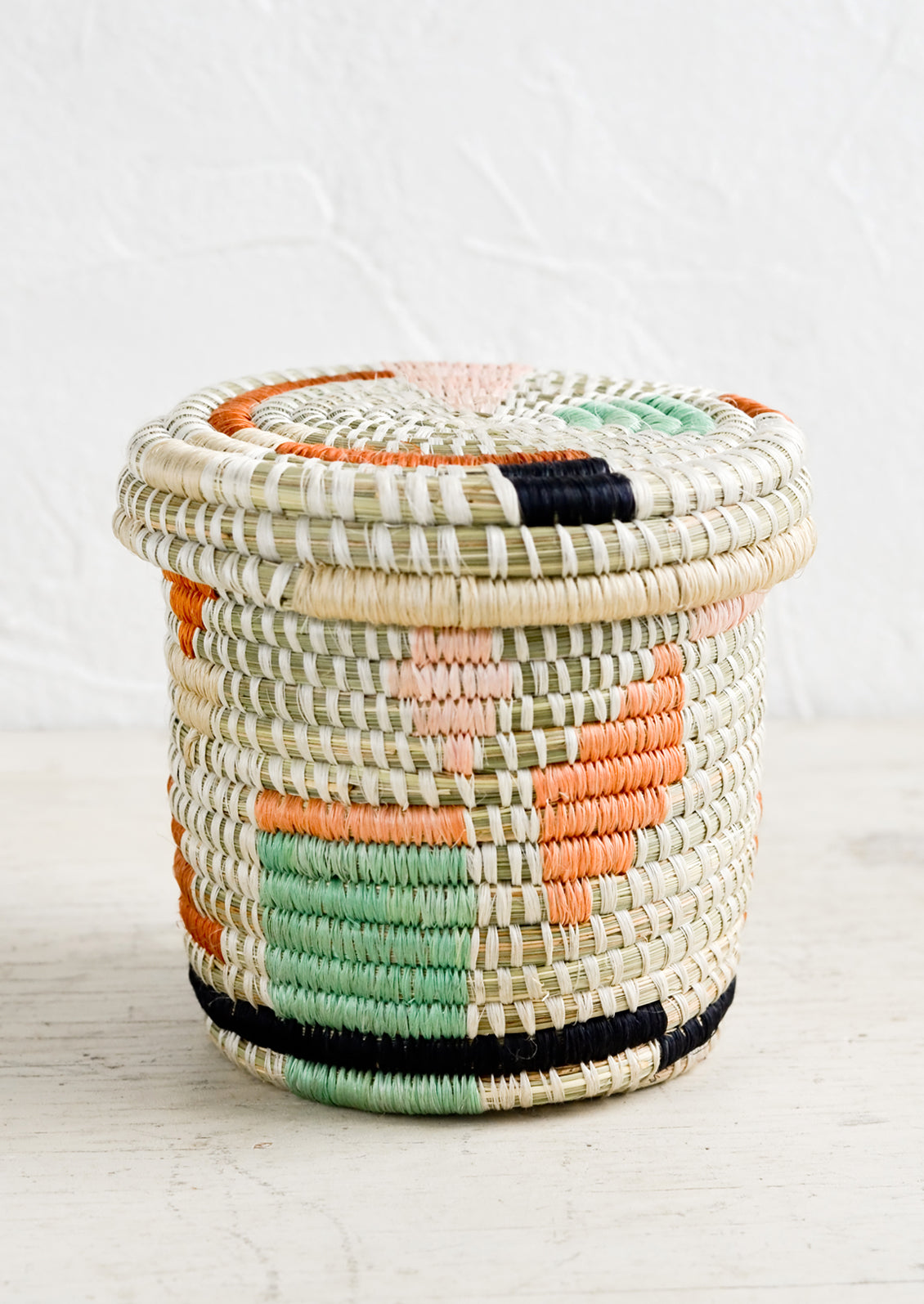 A small, round lidded basket made from woven sweetgrass with geometric pastel pattern.