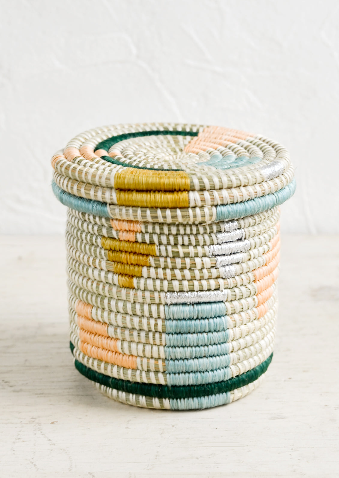 A small, round lidded basket made from woven sweetgrass with geometric pastel and metallic pattern.