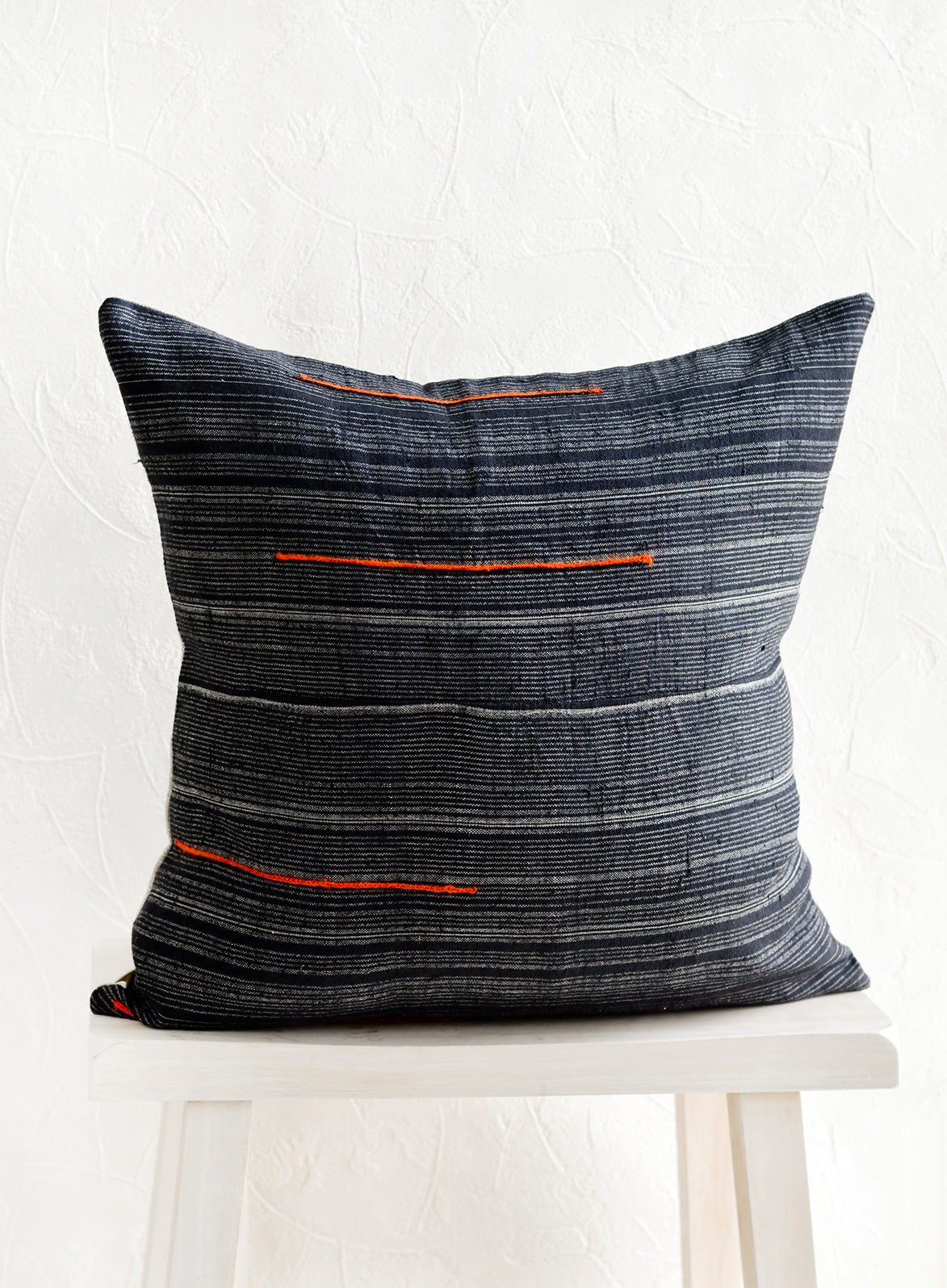 A square throw pillow made from dark blue fabric with grey stripes and embroidered orange lines.