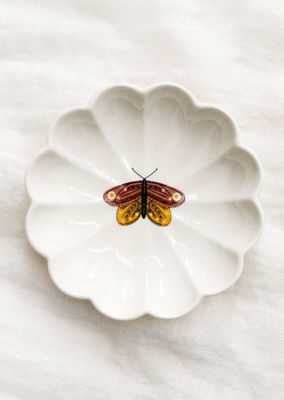 A white ceramic dish with scalloped shape and pink and yellow moth illustration at center.