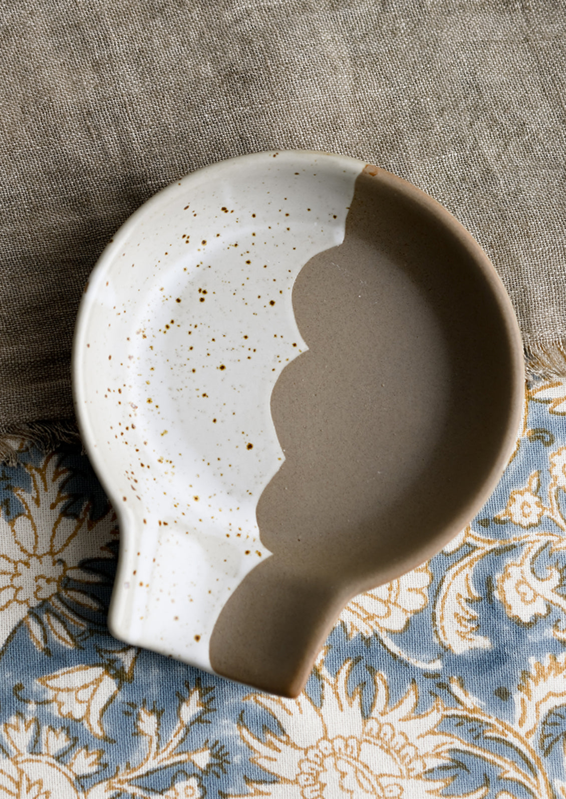 A ceramic spoon rest with scalloped edge dividing brown and speckled white halves.