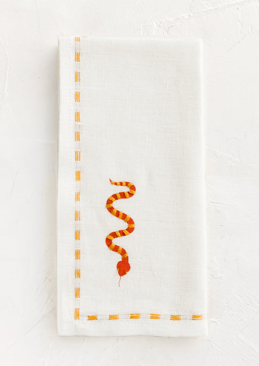 A white linen napkin with yellow stitching and red and yellow snake embroidery at corner.