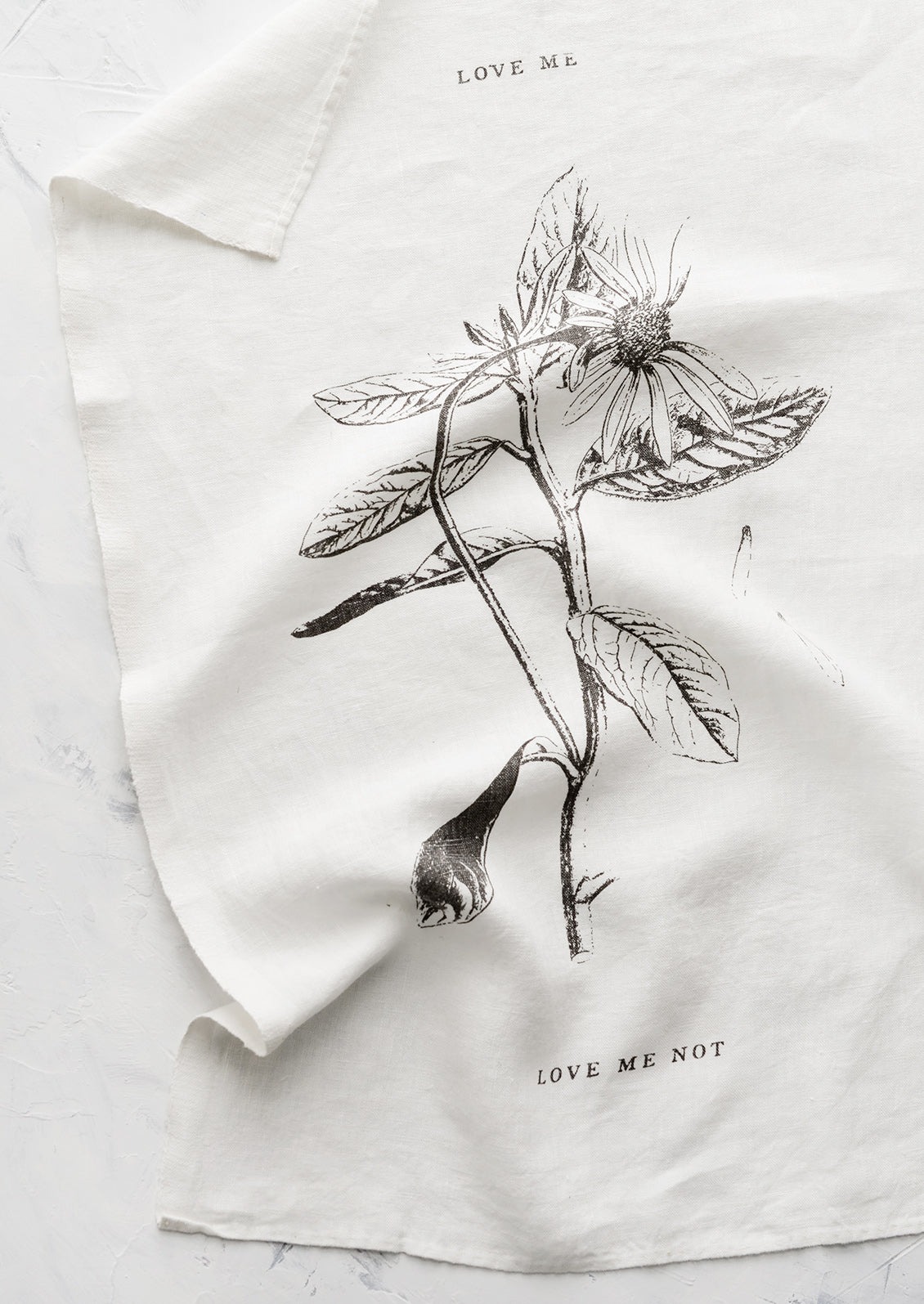 A white linen tea towel with floral image and text reading "love me" and "love me not".