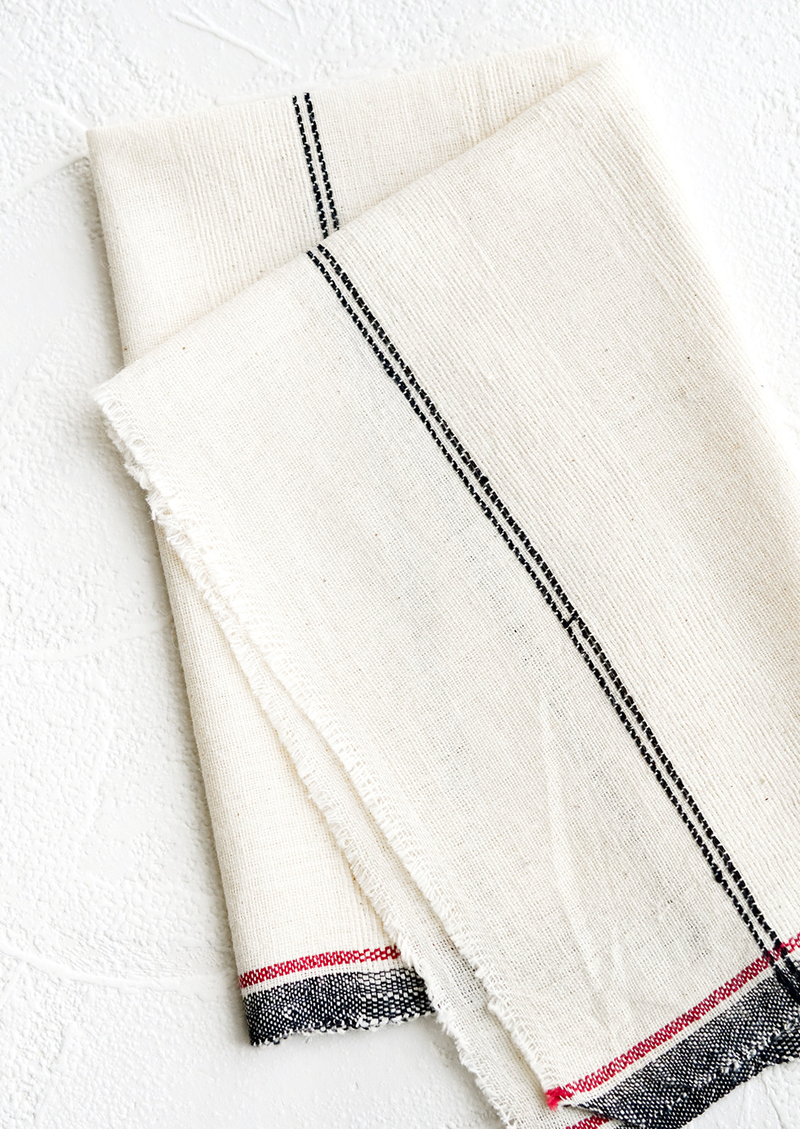 A natural cotton napkin with black and red stripe detailing and raw edges.