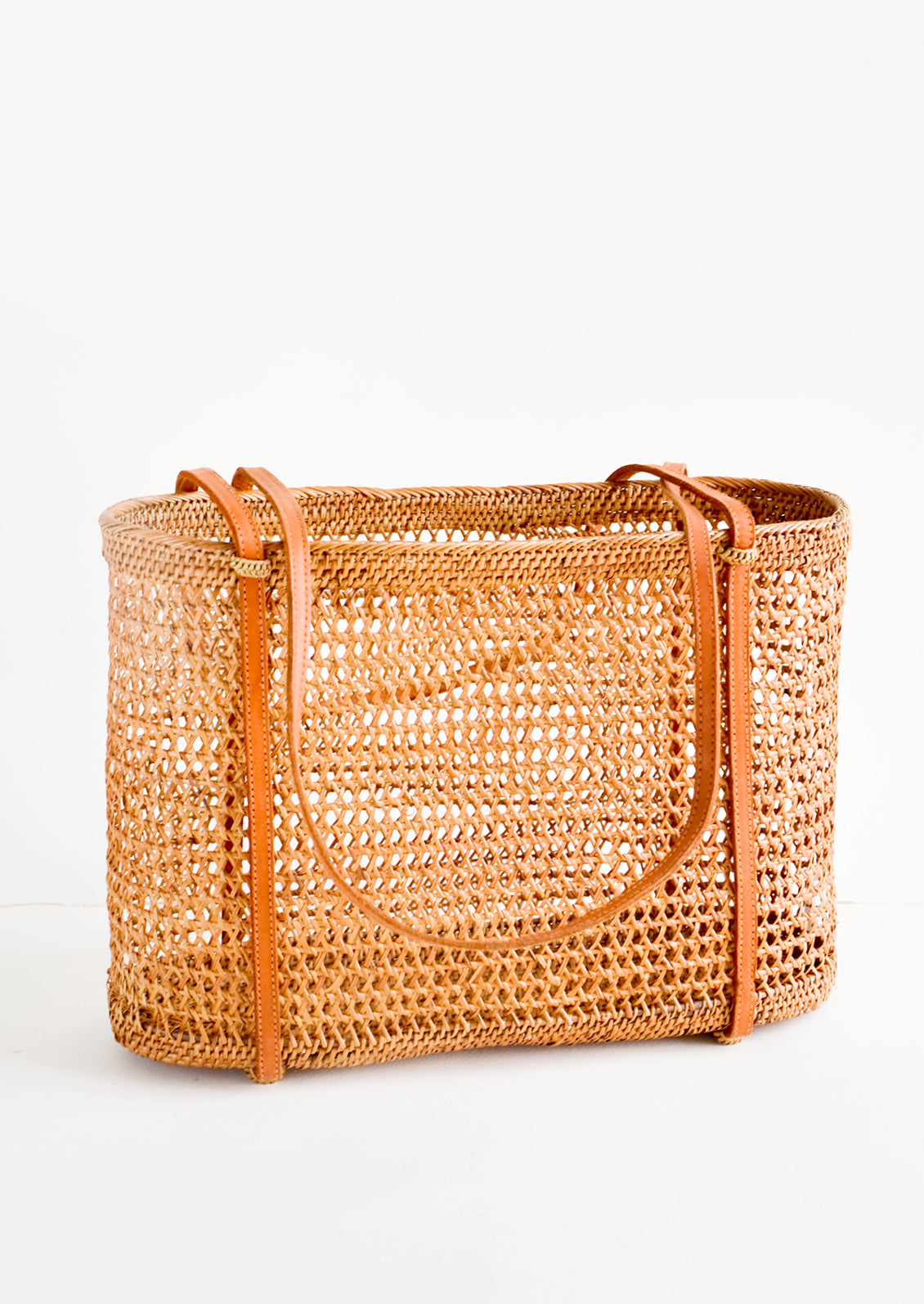 East-west tote bag made from open weave rattan, thin, tanned leather shoulder straps wrap around the bottom of the bag
