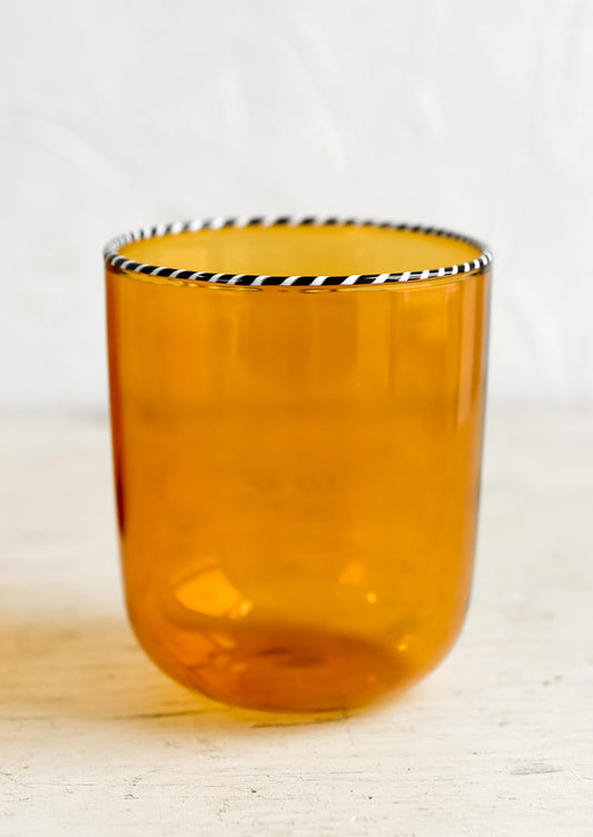 A transparent glass tumbler in amber color with black and white striped rim.