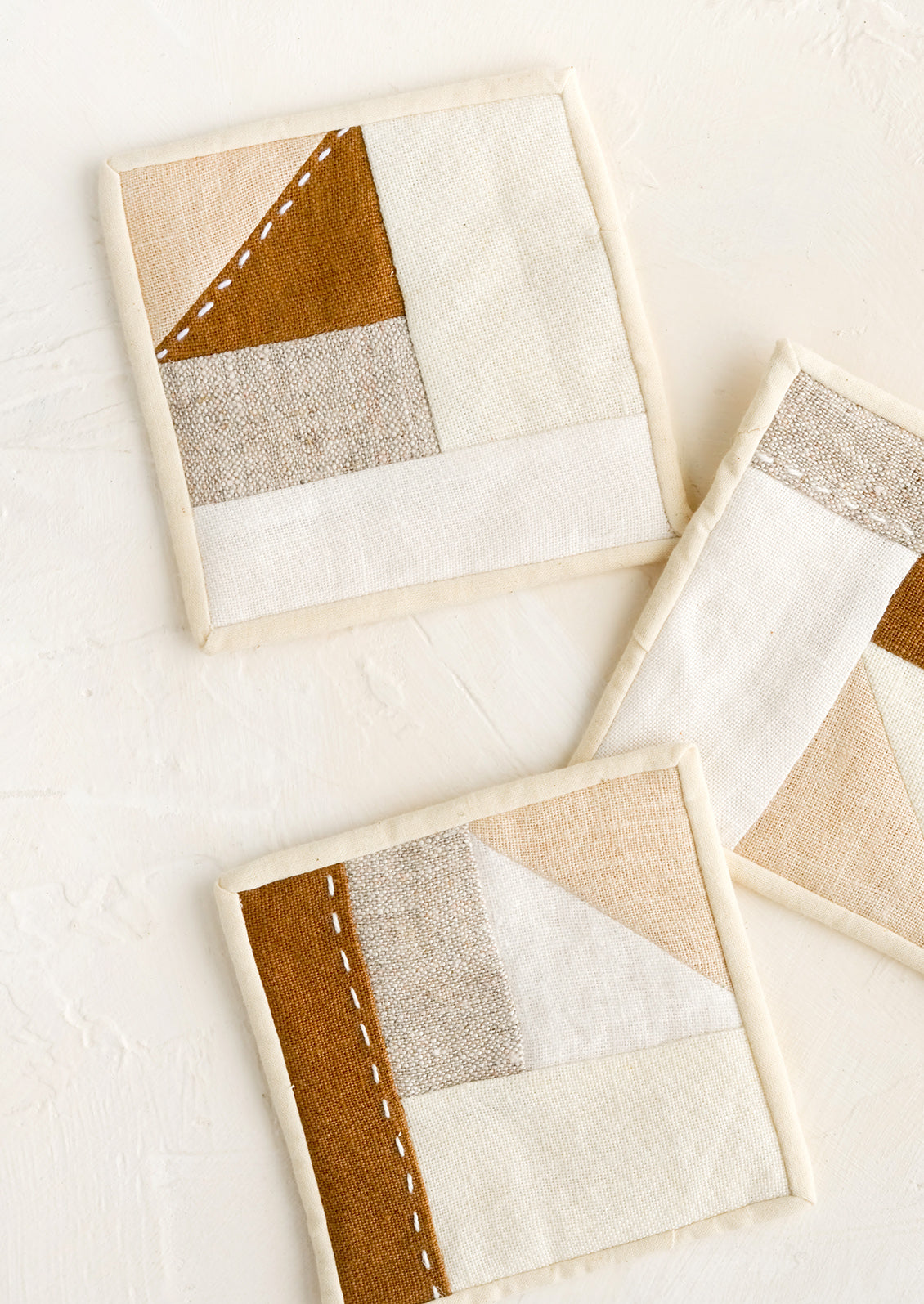 Three square drink coasters made from neutral hued patchwork fabric.