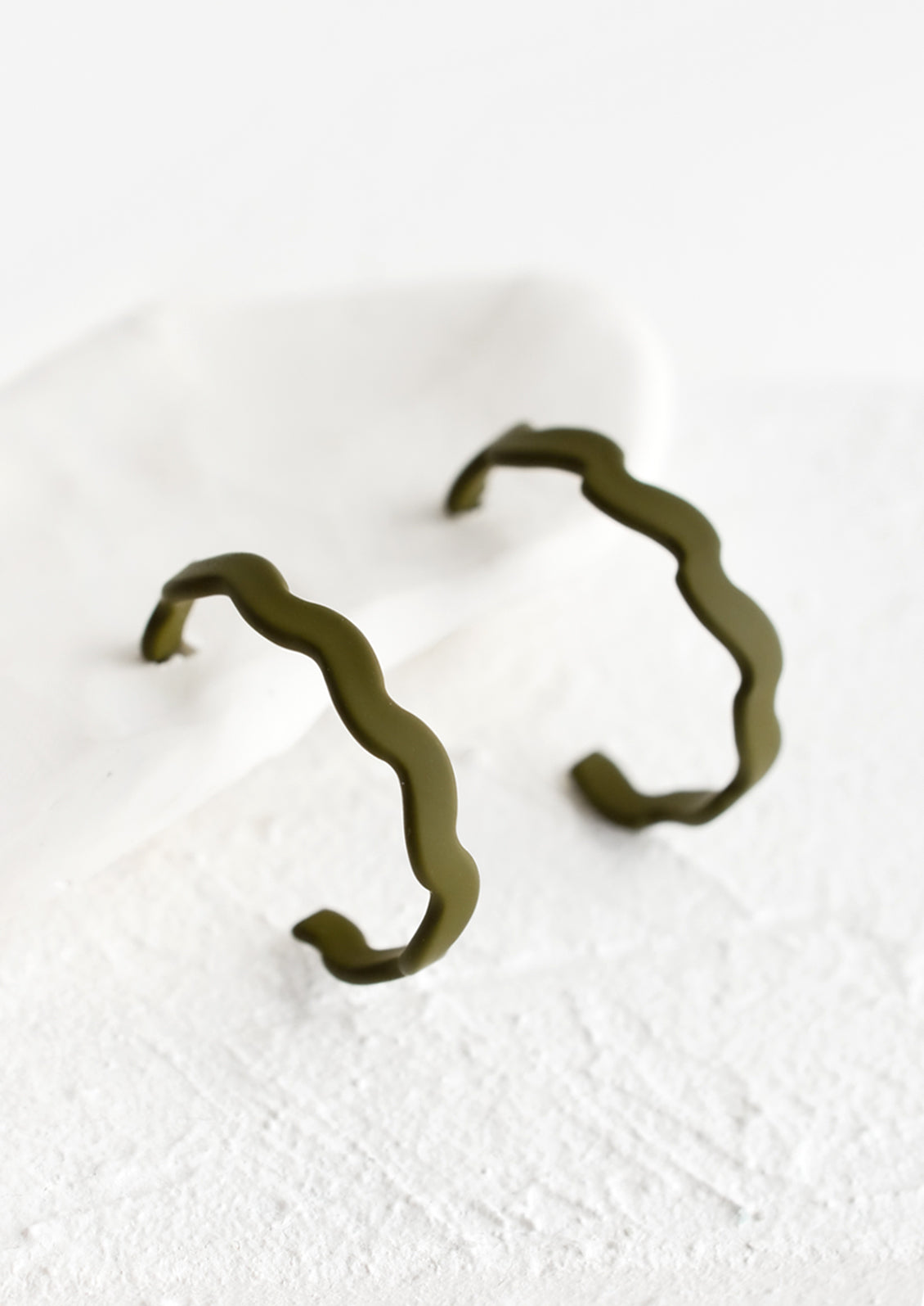 A pair of hoop earrings in matte olive with wavy shape.